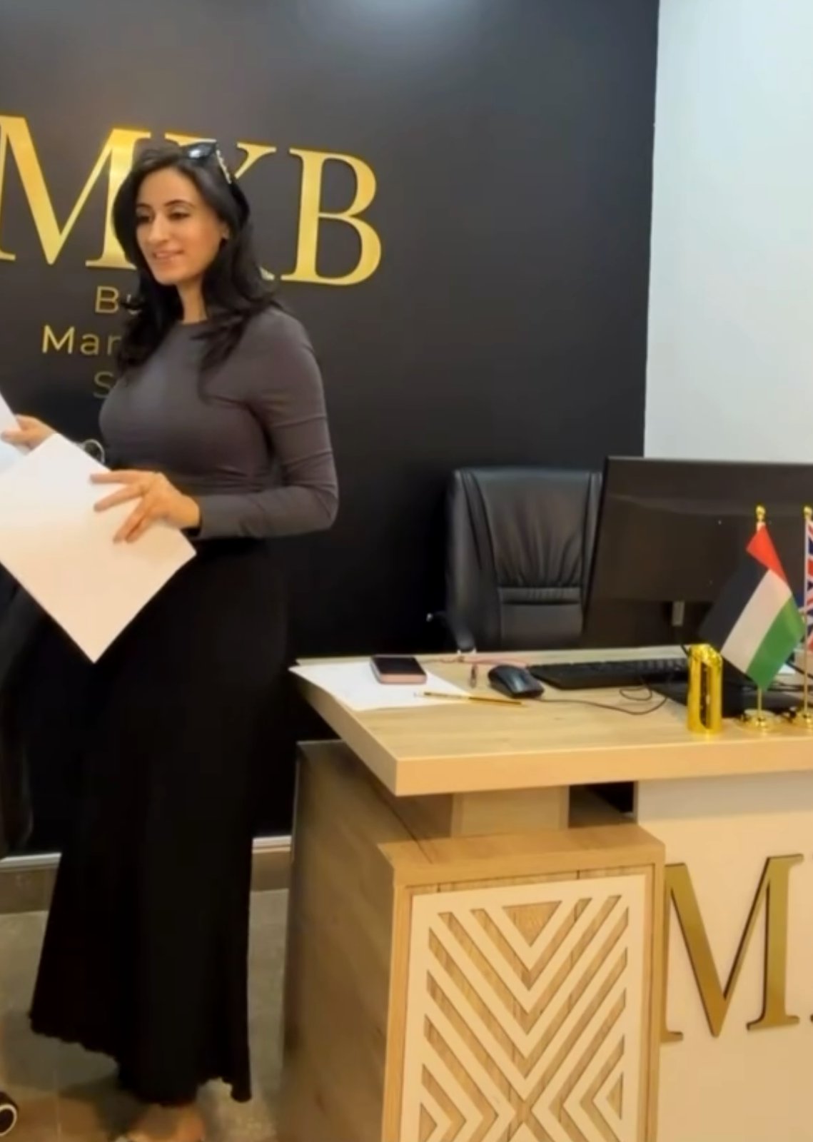 Mehak Butt smiling with client paperwork in front of the gold MKB logo wall